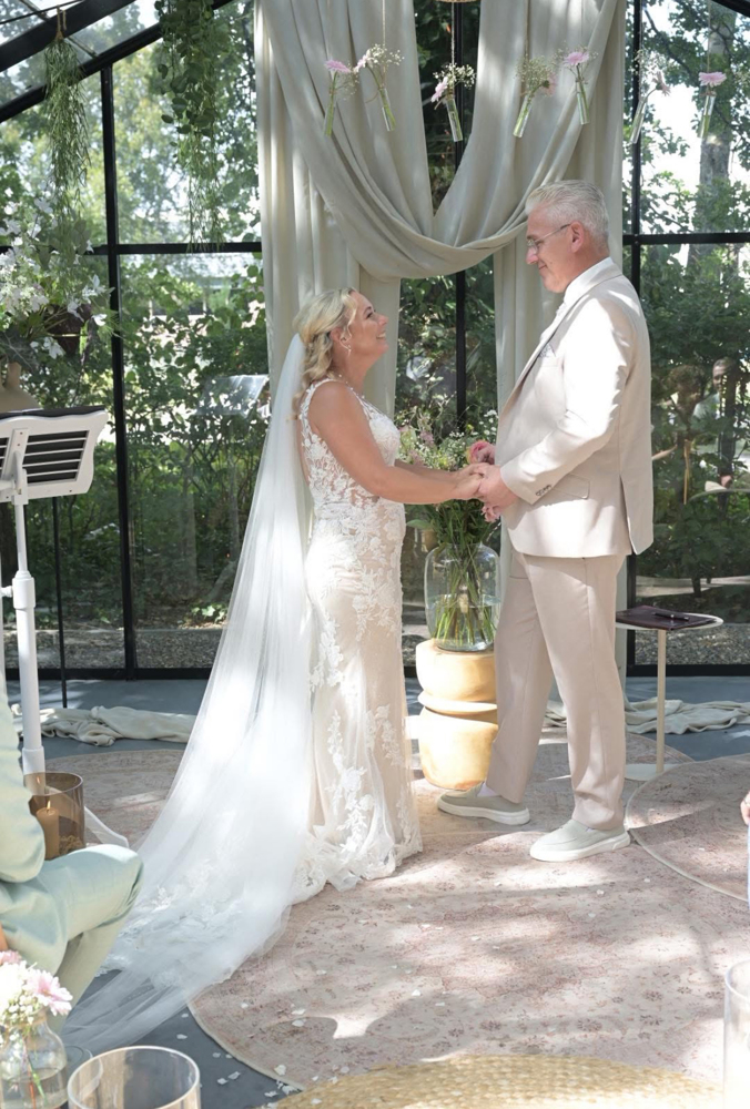 real bride cindy van der putten – elegante fit & flare trouwjurk met lange sluier – bruid en bruidegom geven elkaar de handen tijdens de ceremonie; zacht licht valt door de glazen kas vol groen en bloemen